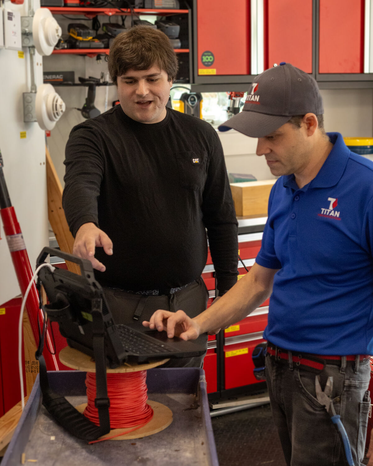 Sam Zimmerman points at a rugged laptop on a cart while Joe Montuori, in his work polo and cap, types on it, in front of a wall of fire alarm devices