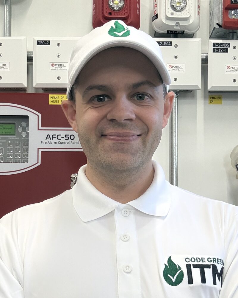 Joe Montuori in a white CodeGreen ITM polo and cap, arms crossed, in front of a fire alarm control panel and a wall of devices